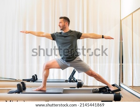 Fit man performing a lunge and stretch Warrior 2 yoga pose on a pilates reformer bed to stretch and strengthen his hip and chest muscles and promote stamina Royalty-Free Stock Photo #2145441869