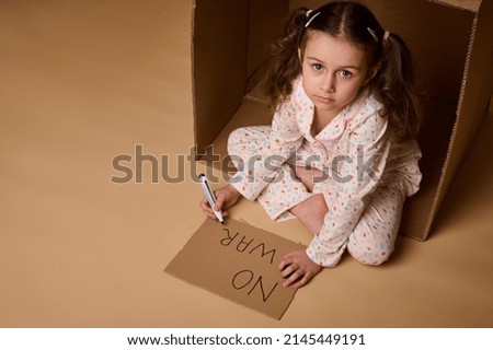 Little girl child in pajamas writing No War on poster while sitting inside a cardboard box, hiding from military and political conflict. Concept of refugees, immigrants losing their home during war