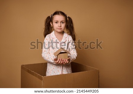 Little Caucasian girl, adorable child in pajamas holds a craft cardboard house model being inside a box, against beige background with copy space for ads. The concept of investment, housing
