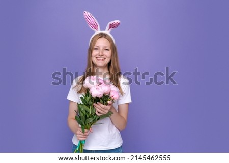 Cute young woman with pink easter bunny ears on purple background in white t-shirt. Caucasian girl holding a bouquet of pink peonies. Waiting for a bright spring holiday