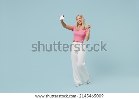 Full body young blonde woman 20s in casual pink t-shirt doing selfie shot on mobile cell phone post photo on social network show v-sign isolated on plain pastel light blue background studio portrait.