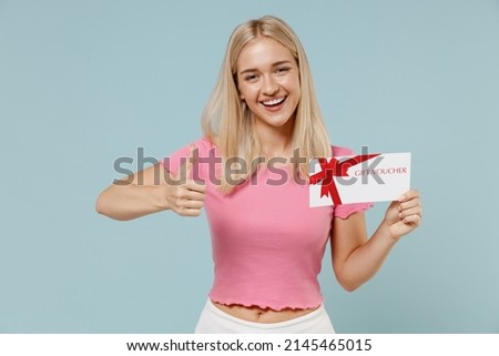 Fun young blonde woman 20s she wears casual pink t-shirt hold in hand mock up of gift certificate coupon voucher card for store show thumb up like isolated on plain pastel light blue background studio