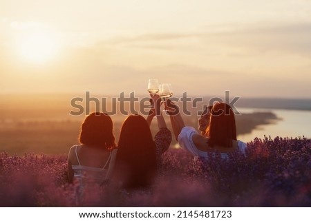 Back view of company of young female friends with hands up having fun, raising glasses with wine and enjoy beautiful sunset at summer picnic in lavender field. Royalty-Free Stock Photo #2145481723