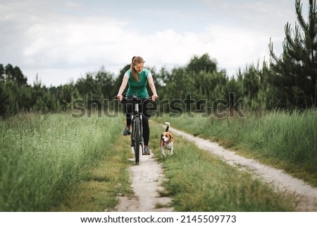 Woman cycling with a dog. Young woman riding bicycle together with her beagle dog pet running nearby. Traveling with a dog Royalty-Free Stock Photo #2145509773