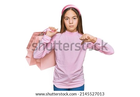 Beautiful brunette little girl holding shopping bags with angry face, negative sign showing dislike with thumbs down, rejection concept 