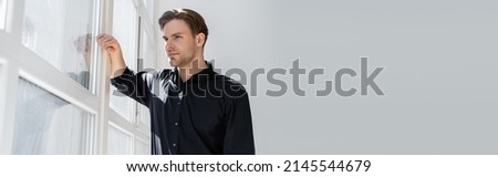 pensive man in black shirt standing near window and looking away, banner