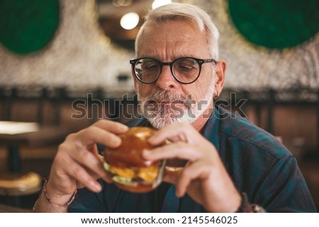 Senior gray-haired man in glasses with a beard is eating an appetizing burger. Lunch break, rest in the pub.