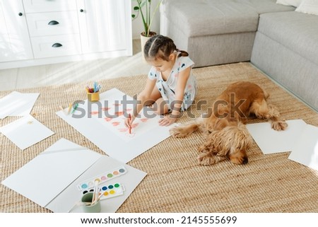 A girl draws hearts for his mother sitting on carpet floor in living room, cocker spaniel dog lying nearby