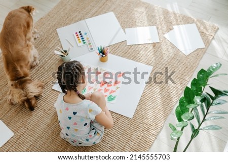 A girl draws hearts for his mother sitting on carpet floor in living room, cocker spaniel dog sitting nearby