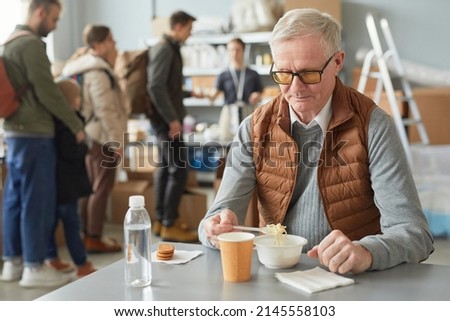 Portrait of Caucasian senior man eating simple meal at soup kitchen in help center for refugees, copy space Royalty-Free Stock Photo #2145558103