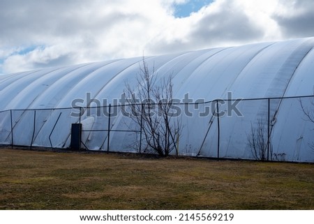 The exterior of a white semi permanent indoor tennis court dome tent. The polythene symmetrical pattern of the prefabricated pressurized air bubble is next to a grassy field and wire fence. Royalty-Free Stock Photo #2145569219