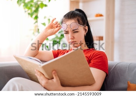 A young brunette with glasses is reading a book, having poor eyesight at home. a student experiencing vision problems or eye fatigue is reading a book. Royalty-Free Stock Photo #2145593009