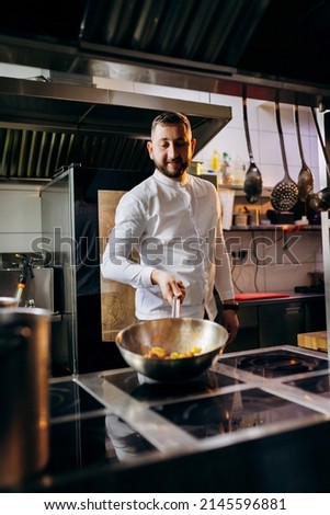 Сhef in the professional kitchen with a frying pan and a fire.
Chef's hands hold iron Pan and preparing food on cooker.