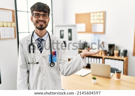 Hispanic man with beard wearing doctor uniform and stethoscope at the office smiling cheerful presenting and pointing with palm of hand looking at the camera. 