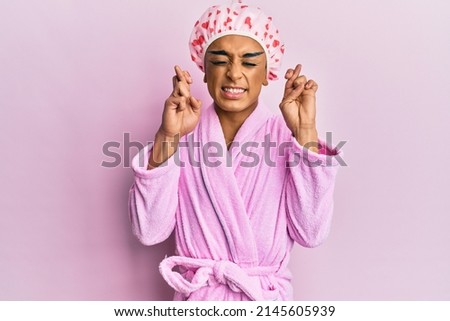 Hispanic man wearing make up wearing shower towel cap and bathrobe gesturing finger crossed smiling with hope and eyes closed. luck and superstitious concept. 