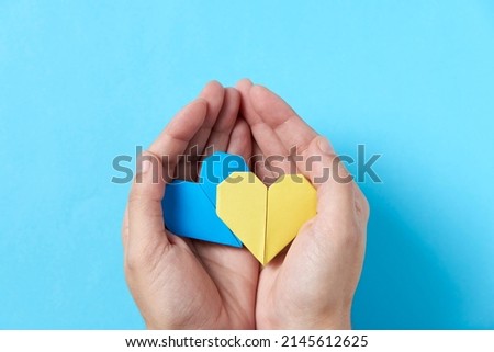 Hands of an unrecognizable person holding and protecting a pair of blue and yellow origami hearts, colors of the Ukrainian flag. Solidarity with Ukraine during the 2022 Russian military invasion.