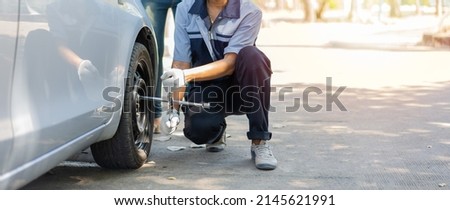 Expertise mechanic man  in uniform using force trying to unscrew the wheel bolts nuts and help a woman for changing car wheel on the highway, car service, repair, maintenance concept. Royalty-Free Stock Photo #2145621991