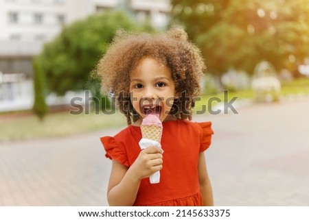a black child eats ice cream on a hot day. Ice cream in a waffle cone. A happy and contented child in a red dress at summer.