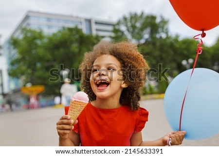 A black laughing child in a red dress eats ice cream on a hot day. Ice cream in a waffle cone. A happy and contented child at summer.