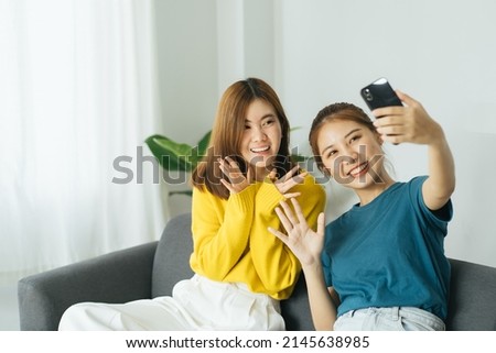Two Pretty young asian female with big smile sitting at living room. She having fun taking light cheerful selfie on blurred background.