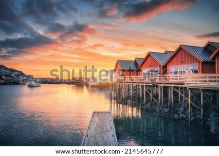 Red rorbu on wooden piles on sea coast, small jetty, colorful orange sky at sunrise in winter. Lofoten islands, Norway. Traditional norwegian rorbuer, reflection in water, boats in fishing village Royalty-Free Stock Photo #2145645777