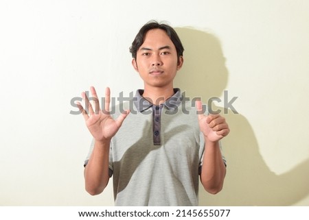 asian man posing smiling and looking at camera showing six (6) fingers. Portrait of Indonesian man in gray t-shirt on white background isolated.
