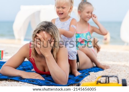 Two cute little sibling girls enjoy having fun playing sitting on tired exhausted mother's back at sea ocean beach. Frustrated mom make face palm gesture. Vacation family small kids trouble concept Royalty-Free Stock Photo #2145656005