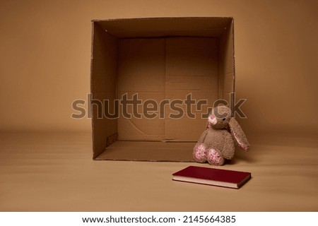 A plush rabbit toy and book next to an empty cardboard box, isolated over beige background with copy ad space