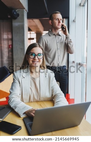 Cheerful female manager in a jacket doing office work and a smiling, successful female boss in optical glasses for vision correction posing at a desk. In the background a man with a phone