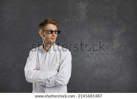 Confident young man in thug life glasses standing with his arms folded and looking away at some copy space. Portrait of funny guy in meme sunglasses against grey background with blank text copyspace