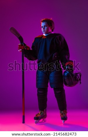 Portrait of little boy, child, hockey player in special uniform posing with stick isolated over purple background in neon light. Concept of action, sport, health, hobby, childhood, team sport