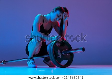 Rest time. Studio shot of female athlete, sportive woman posing with barbell isolated on purple background in neon light. Sport, beauty, strength concept. Female sports