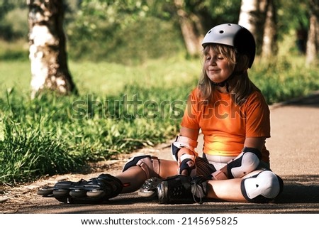 Happy child resting after playing sports on a sunny day. A girl dressed in a sports uniform and roller skates sits on a path in the park on a summer day.