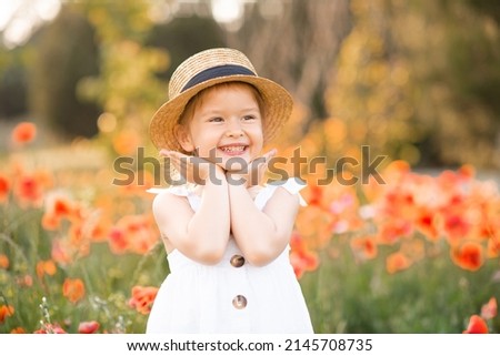 Cute funny baby girl 2-3 year old wear straw hat and white summer dress over flower meadow background. Kid laughing outdoors over nature. Looking at camera. Childhood. 