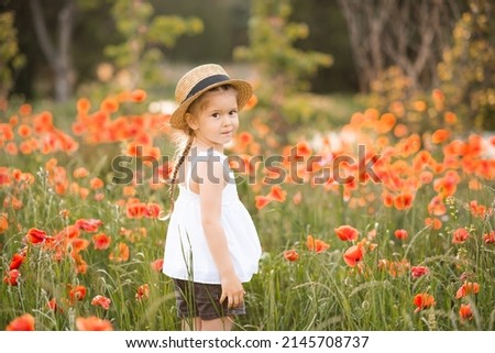 Smiling baby girl 3-4 year old wear straw hat and white rustic dress posing over nature poppy field background. Happy kid in flower meadow. Spring time. Childhood. 