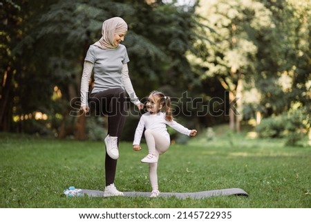 Yoga and stretching exercises of mom with child outdoors. Likable young Muslim mother and her cute daughter, having outdoor workout in the park, standing on yoga mat and doing high stepping exercise Royalty-Free Stock Photo #2145722935