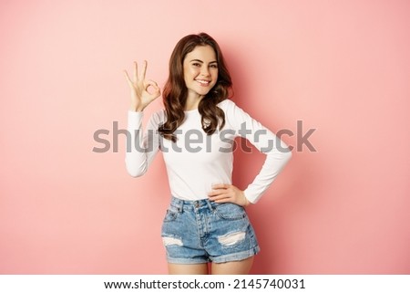 Stylish glamour girl with dark curly hairstyle, showing okay gesture, ok satisfied sign, smiling pleased, complimenting, recommending something good, pink background