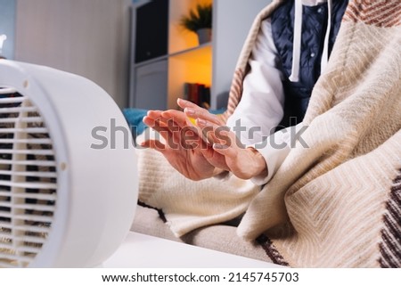 Woman warm of hand from electrical heater in living room without central heating. Energy crisis in Europe Royalty-Free Stock Photo #2145745703