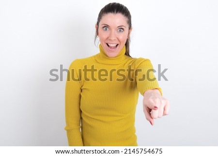 Excited positive Young caucasian woman wearing yellow turltleneck over white background points index finger directly at you, sees something very funny. Wow, amazing