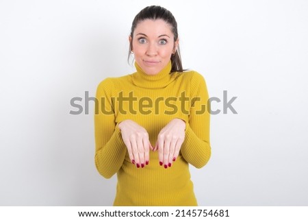 Young caucasian woman wearing yellow turltleneck over white background makes bunny paws and looks with innocent expression plays with her little kid