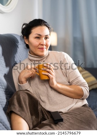 Asian woman sitting on sofa, holding cup of tea and watching television.