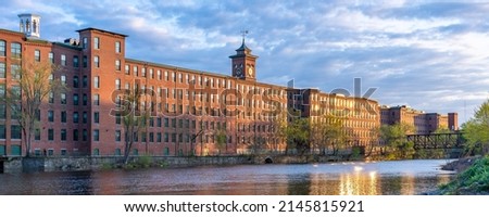 Historic cotton mill building with clock tower in an old industrial park on the Nashua River illuminated by the sun during sunset in May. Panoramic photography. Nashua, New Hampshire, USA