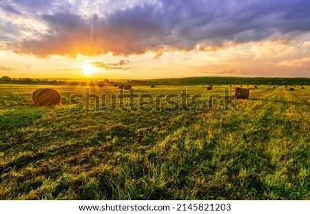 Scenic view at picturesque burning sunset in a green shiny field with hay stacks, bright cloudy sky , trees and golden sun rays, summer valley landscape Royalty-Free Stock Photo #2145821203