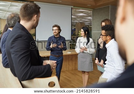 Business coach talking to group of people standing in office around him. Team of entrepreneurs and listening to business trainer speaking about creativity and teamwork, sharing advice and experience Royalty-Free Stock Photo #2145828591