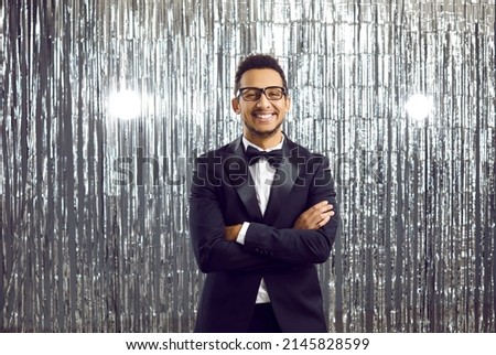 Happy handsome black man in a modern tuxedo, white shirt, bowtie and glasses attending a party. Smiling African gentleman in an elegant suit standing with his arms crossed on a shiny studio background Royalty-Free Stock Photo #2145828599