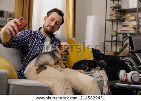 A handsome man in a plaid shirt is sitting on a couch. The dude is taking a selfie with his dogs and wants to put a new post on his social media profiles. A man in the world of social media.