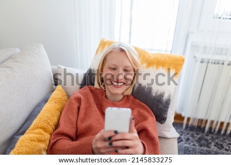 Young woman sit relax on sofa in living room browsing surfing wireless internet on smartphone, millennial girl rest on couch at home message text on modern cellphone, shopping online via website