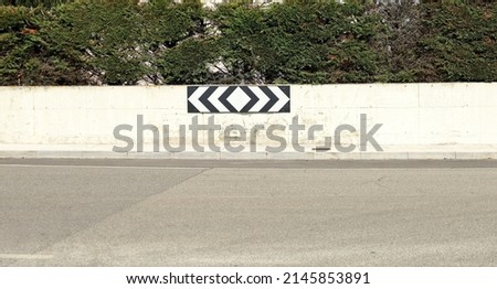 House fence consisting of a concrete wall and a hedge above. A road sign, the roundabout directional arrow,  hanging on the wall. Cement sidewalk and paved street in front. Background for copy space.	