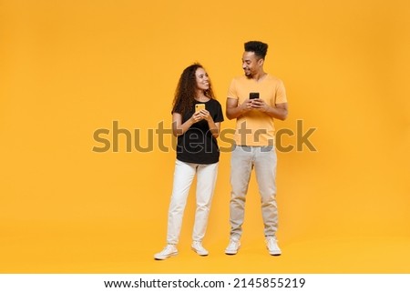 Full length young couple two friends together family african smiling happy woman man in black t-shirt show hold using mobile cell phone look to each other isolated on yellow background studio portrait