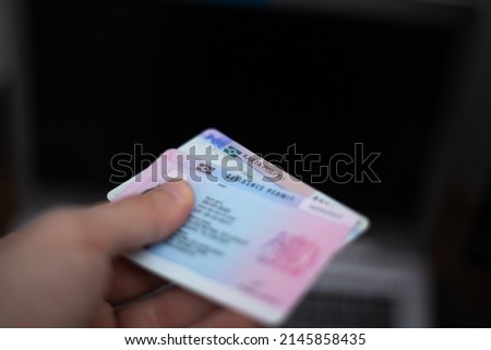 Person holds UK Residence Permit - BRP card and Poland Residence card (Karta Pobytu)  in hand and computer in the background. Immigration concept image. Royalty-Free Stock Photo #2145858435